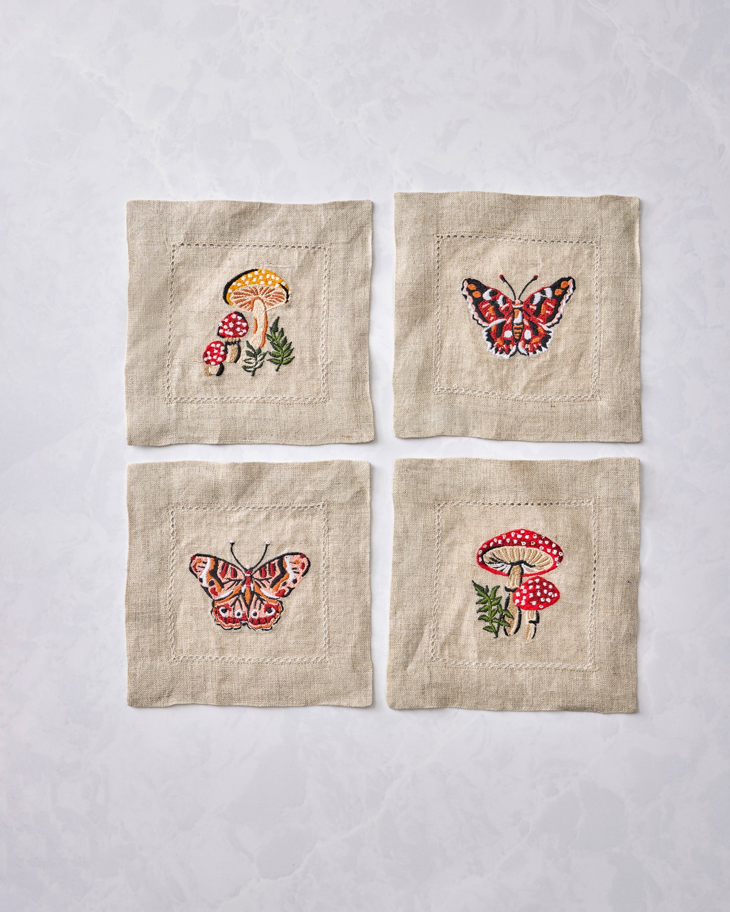Four square embroidered cocktail napkins displayed in a two-by-two arrangement, with mushroom and butterfly designs.