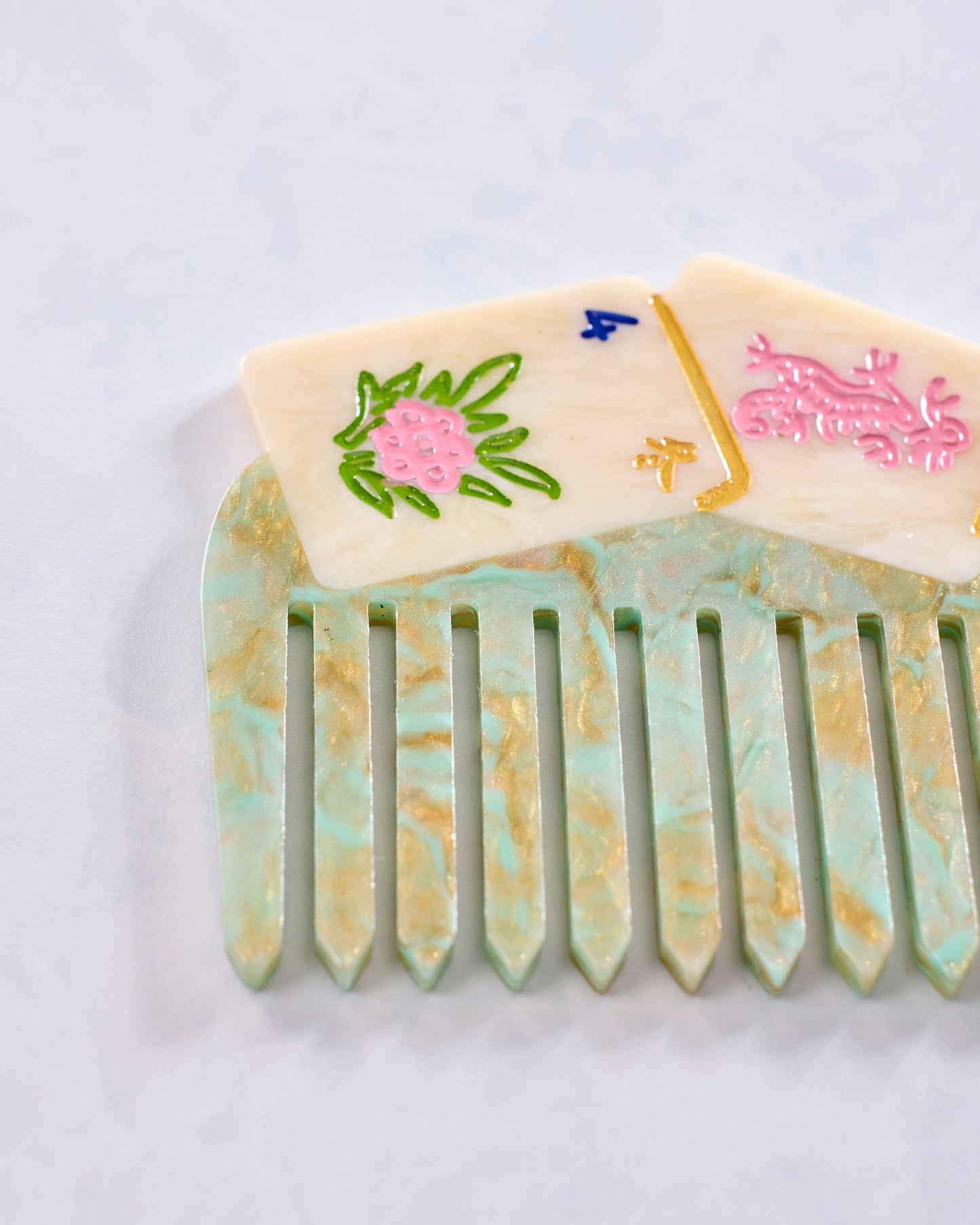 Close-up of a green and cream decorative pocket comb with vertical teeth and painted floral and symbol details.