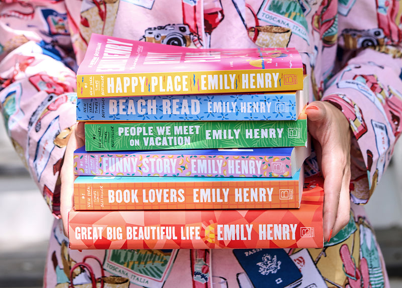 Person in patterned pajamas holds a stack of six Emily Henry books with their spines facing the viewer.