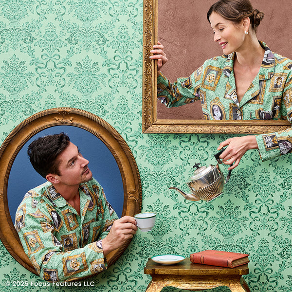 Two people in framed portraits wear patterned pajamas; one pours tea while books sit on a table below.