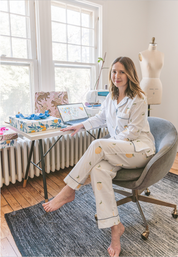 Person seated barefoot in a chair wearing white pajamas with small fruit and plant motifs beside a sewing workspace