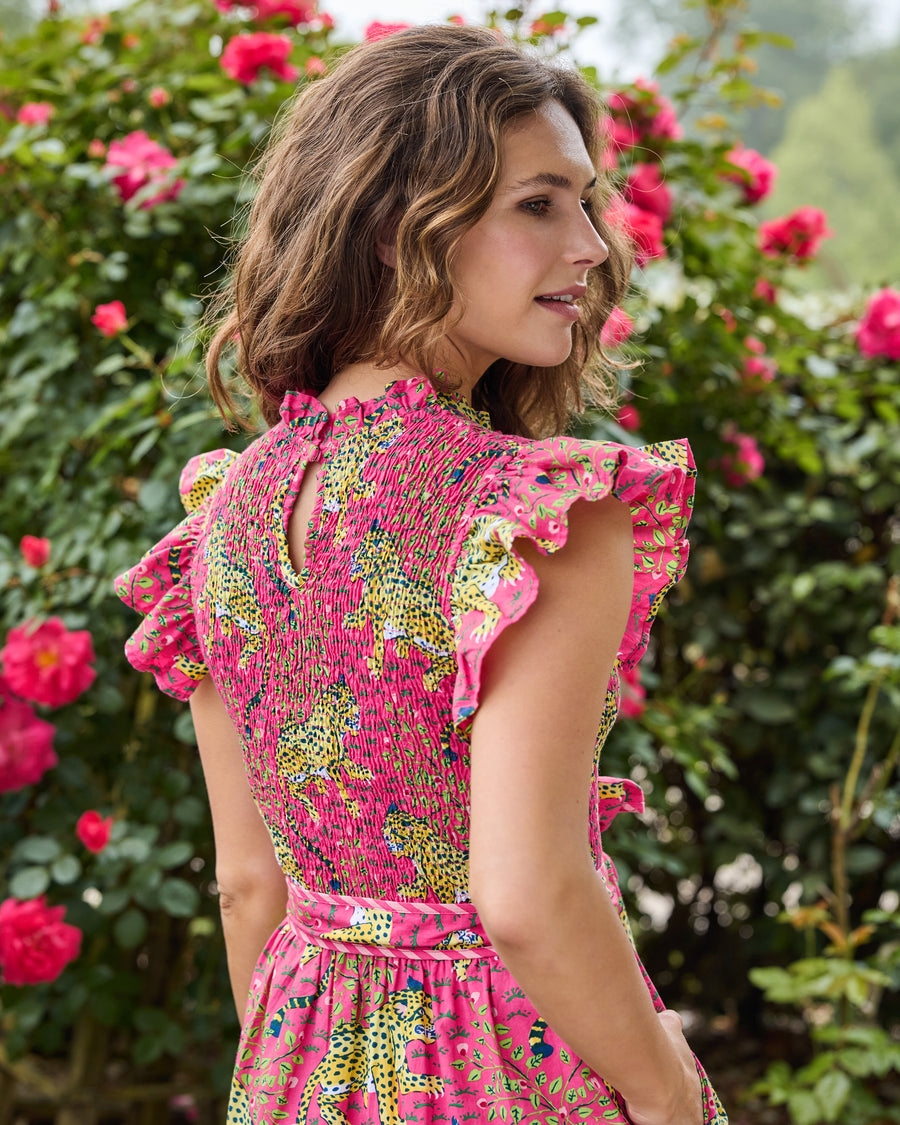Back view of a model wearing a hot pink leopard-print dress with flutter sleeves, a smocked bodice, and a coordinating waist belt in a rose garden.