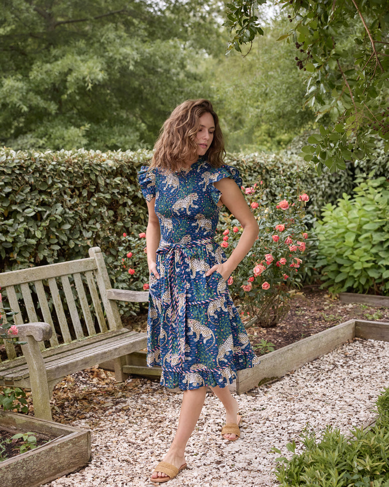 Full-length view of the navy Bagheera Let's Ruffle Dress with leopard print, flutter sleeves, tiered skirt, tied waist belt, and side pockets, worn in a rose garden.