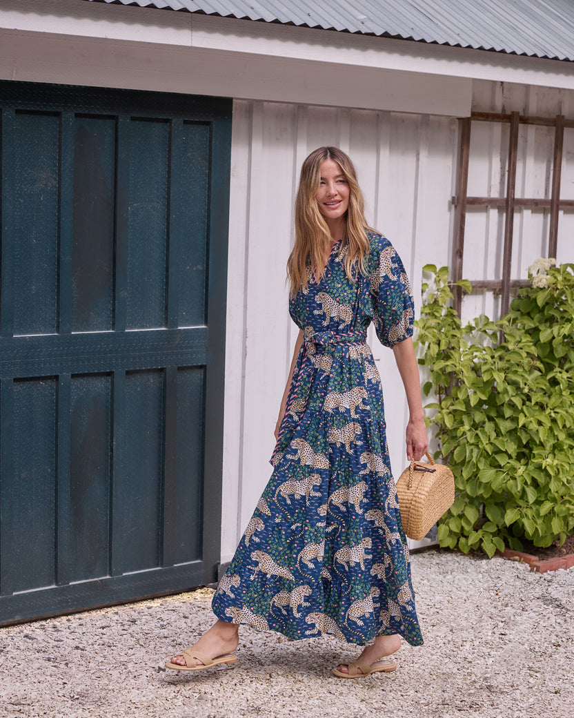 Full-length view of the navy Bagheera dress with a leopard-and-floral print, tied belt, puff sleeves, and a full skirt, worn outdoors with a woven handbag.