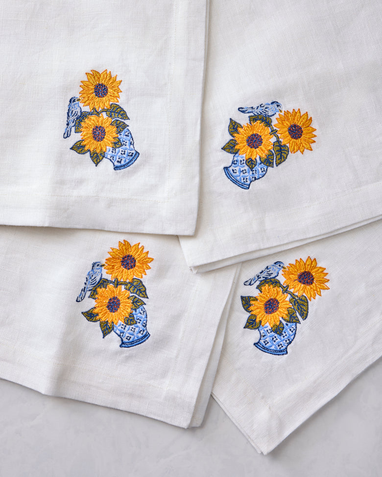 Close-up of four white napkins featuring embroidered sunflowers in blue vases with birds.