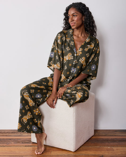 Seated front view of the olive Wildest Dreams pajama set, showing the short-sleeve button-front shirt and wide-leg pants with leopard and flower prints.