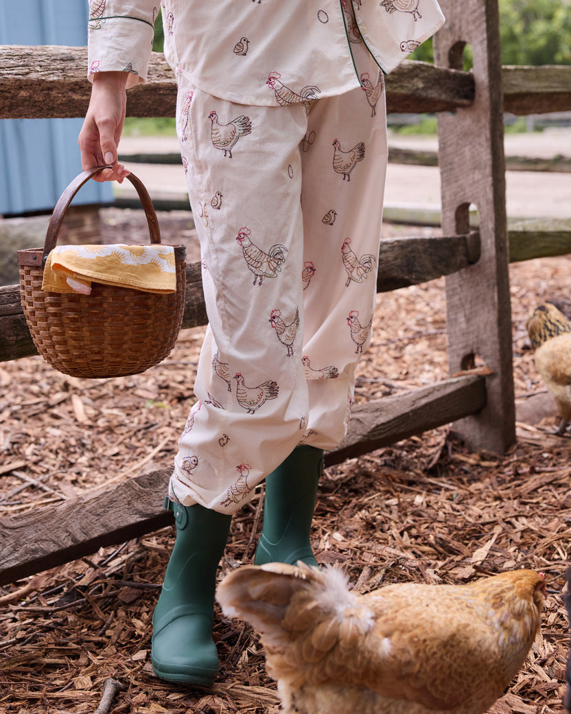 Close view of the buff Embroidered Hens pajama pants with chicken illustrations, shown with green rain boots, a woven basket, and a chicken.
