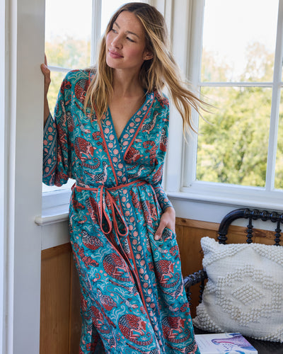 Model wearing the full-length teal Southwestern Critters satin robe beside a window, showing the wrap front, tie belt, wide sleeves, and patterned trim.