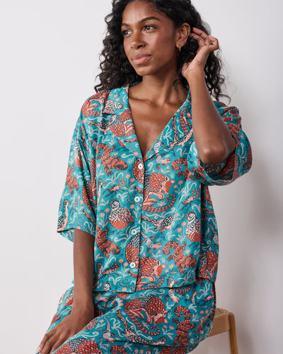 Seated model wearing the teal Southwestern Critters satin Wildest Dreams set, showing a short-sleeve button-front top and matching pants.