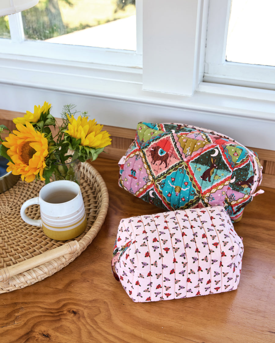 Two quilted oversized pouches with colorful patchwork-style prints beside a tray of sunflowers and a mug.