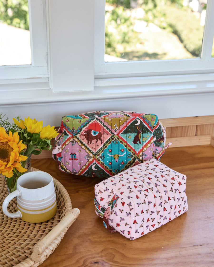 Two quilted oversized pouches with colorful patchwork-style prints on a wooden tabletop beside sunflowers and a mug.
