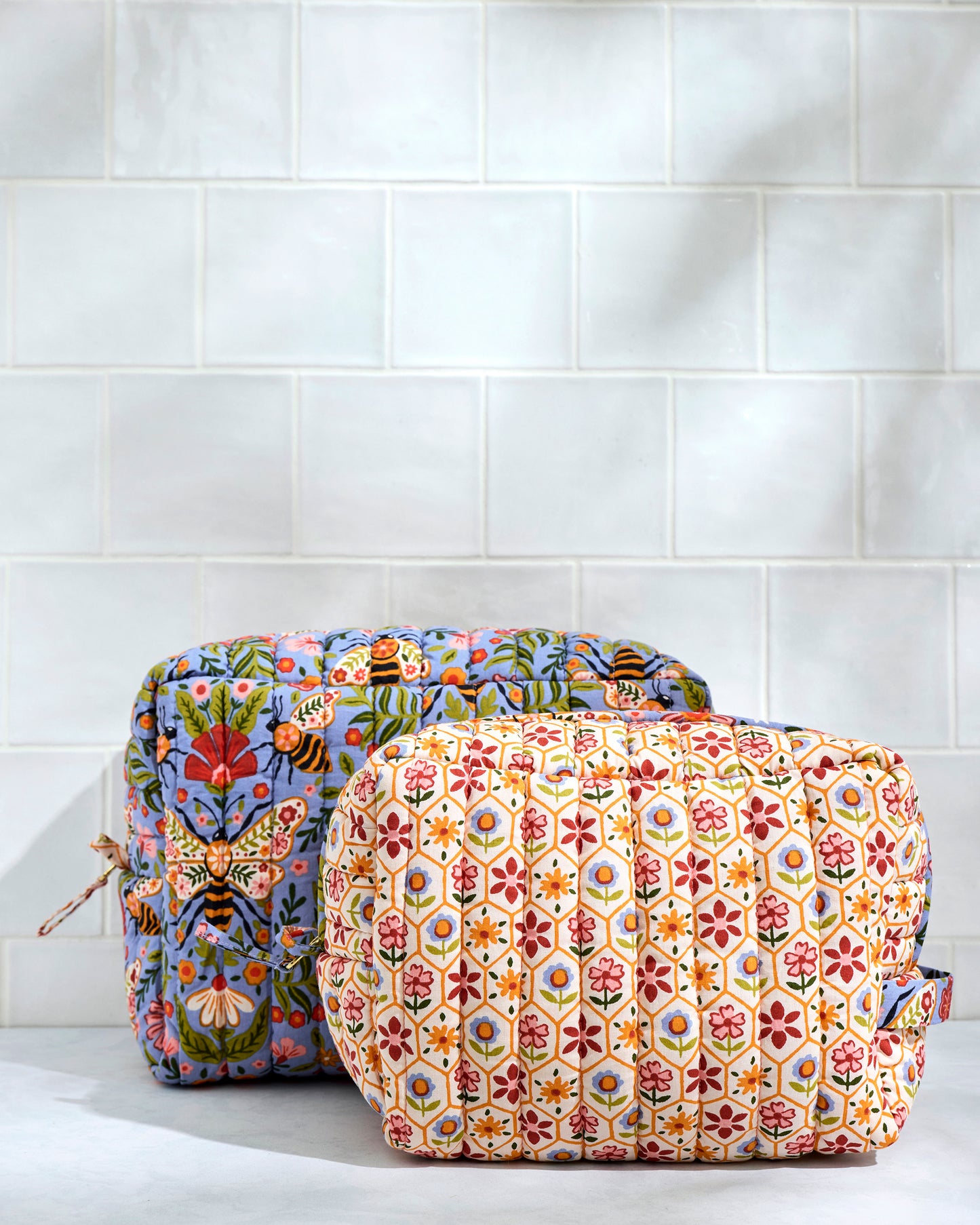 Two closed quilted Bee Tapestry pouches shown from the front, with the cream floral-patterned pouch in front of the blue floral-and-bee pouch.