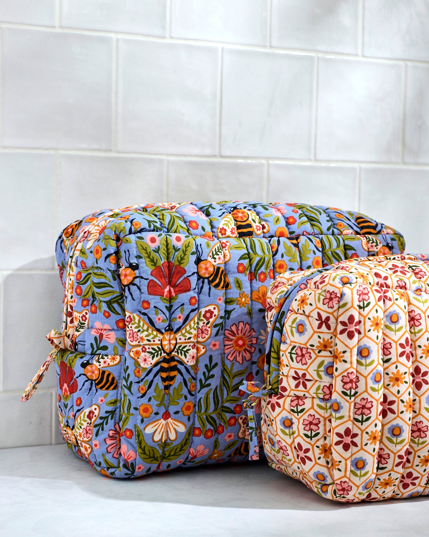 Close view of two quilted Bee Tapestry pouches, showing the blue floral-and-bee pouch behind the cream repeating floral-patterned pouch.