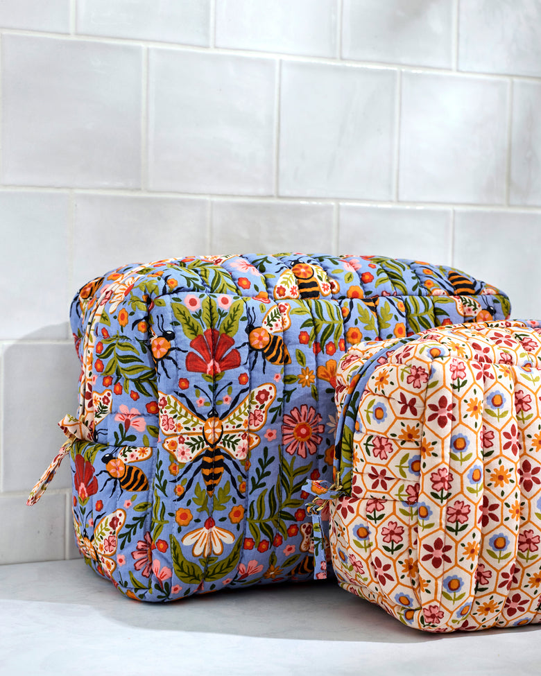 Close view of two quilted Bee Tapestry pouches, showing the blue floral-and-bee pouch behind the cream repeating floral-patterned pouch.