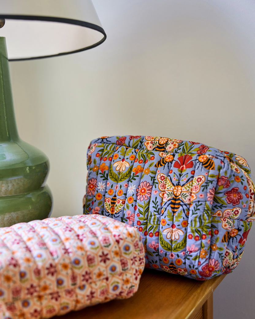 Two quilted Bee Tapestry pouches staged on a wooden table, with the blue floral-and-bee pouch in focus behind a cream patterned pouch.