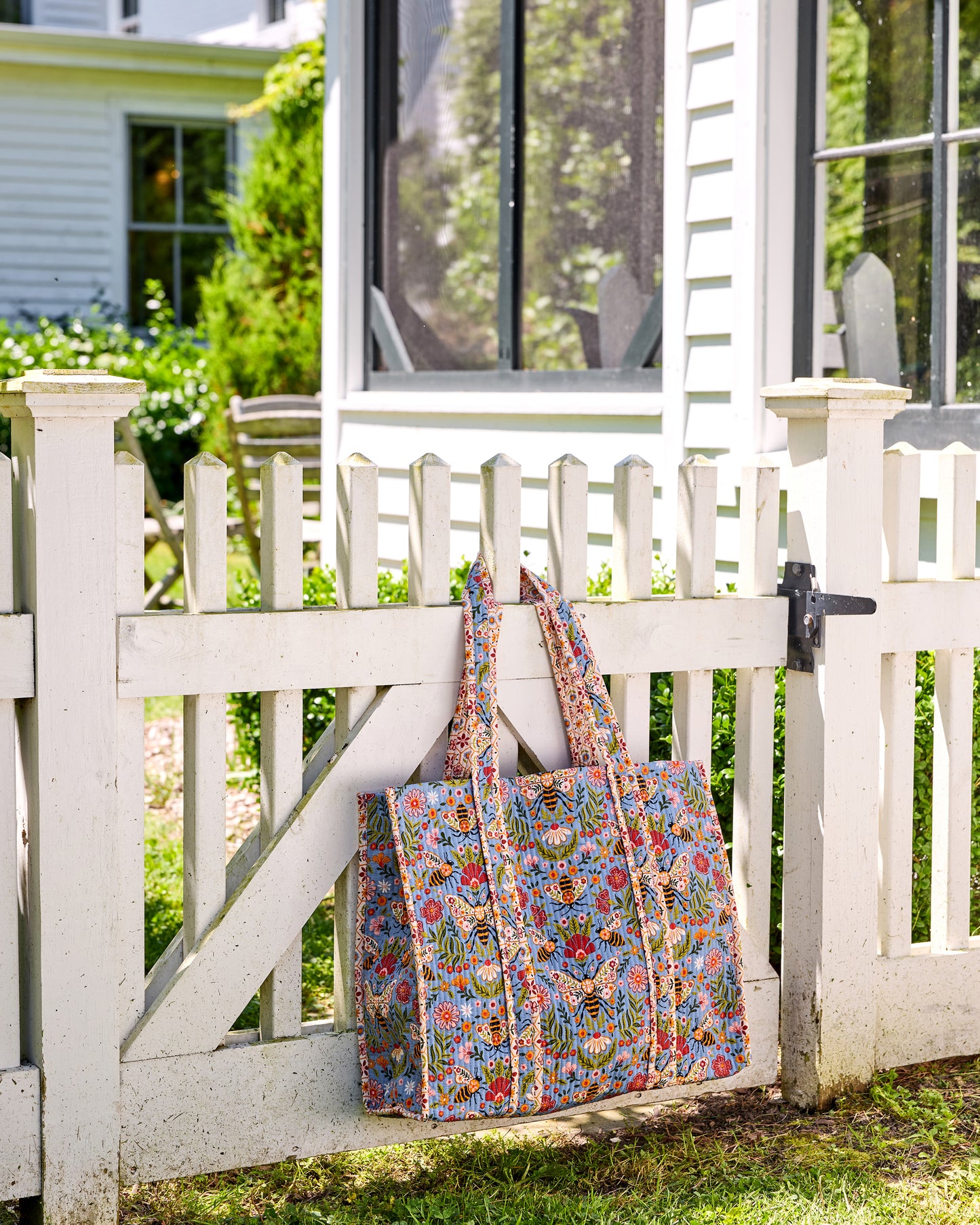 Bee Tapestry traveler tote hanging from a white picket fence, showing its blue bee-and-floral print and long handles.