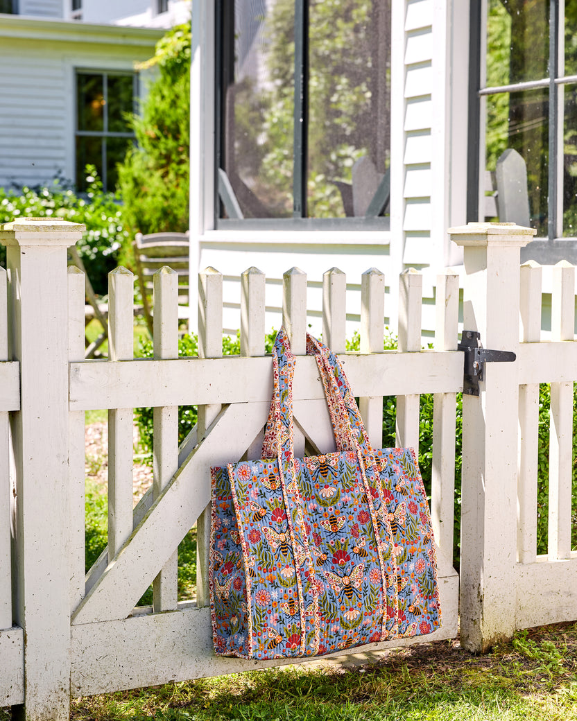 Bee Tapestry traveler tote hanging from a white picket fence, showing its blue bee-and-floral print and long handles.