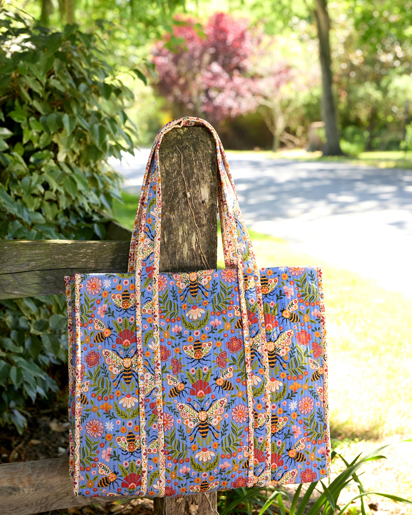 Bee Tapestry traveler tote hanging front-facing on a wooden fence, showing the blue bee-and-floral print and long handles.