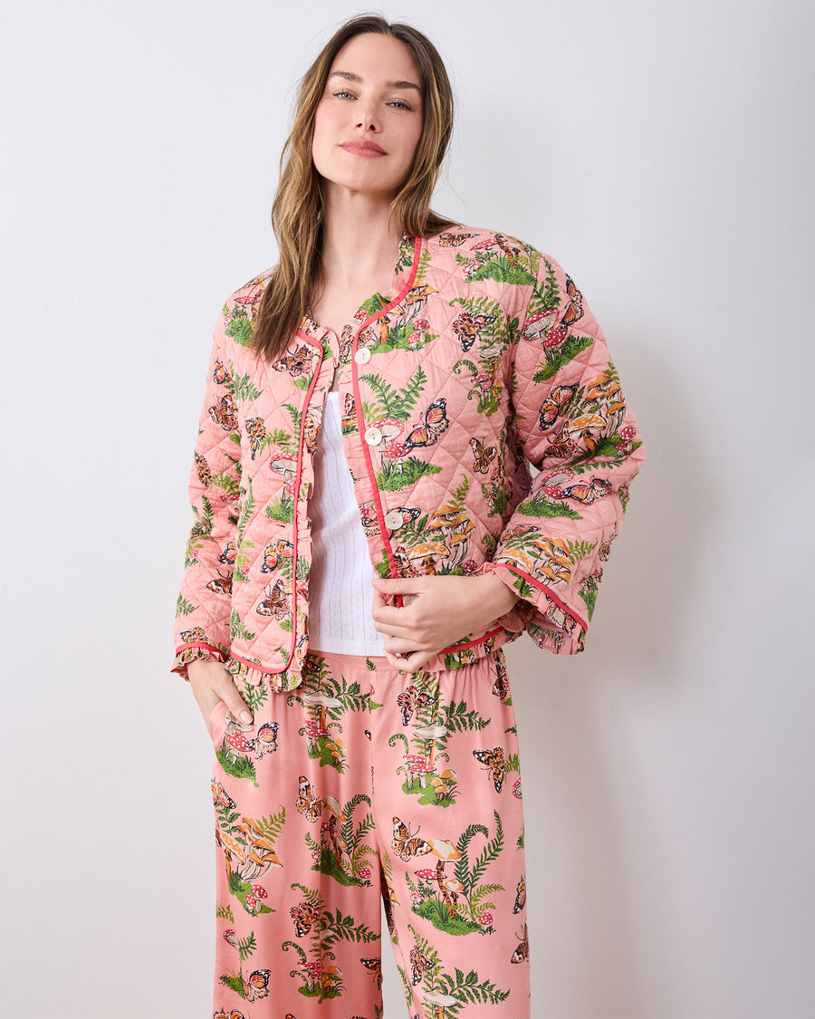 Model wearing the pink Butterfly Forager quilted slumber jacket with ruffle trim over a white top and matching patterned pants.