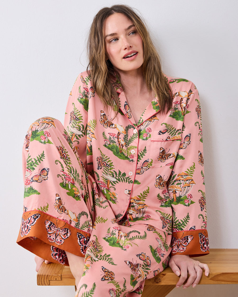 Model seated on a wooden bench wearing the pink Butterfly Forager pajama set with a button-front top, chest pocket, and wide-leg pants.