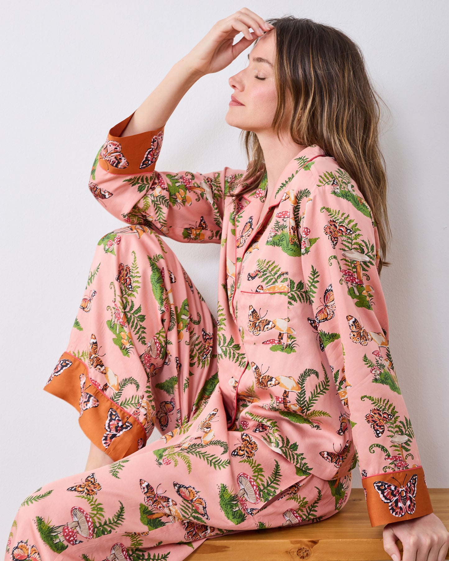 Side view of the pink Butterfly Forager petite satin Soho pajama set worn while seated on a wooden bench.
