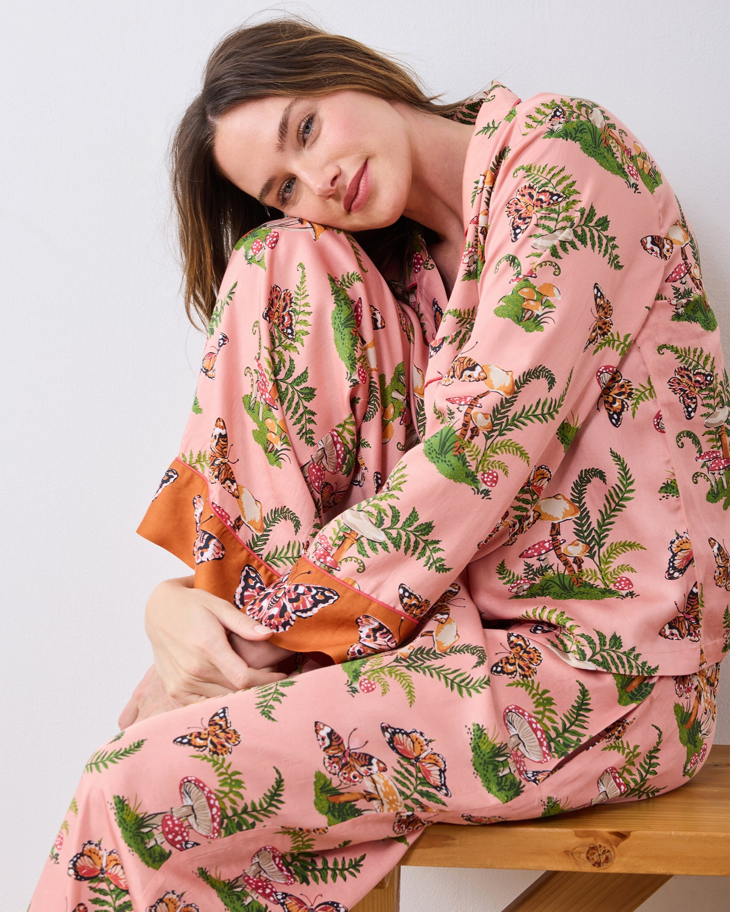 Close view of a model seated on a wooden bench wearing the pink Butterfly Forager tall pajama set, showing the patterned top, pants, and contrasting cuffs.