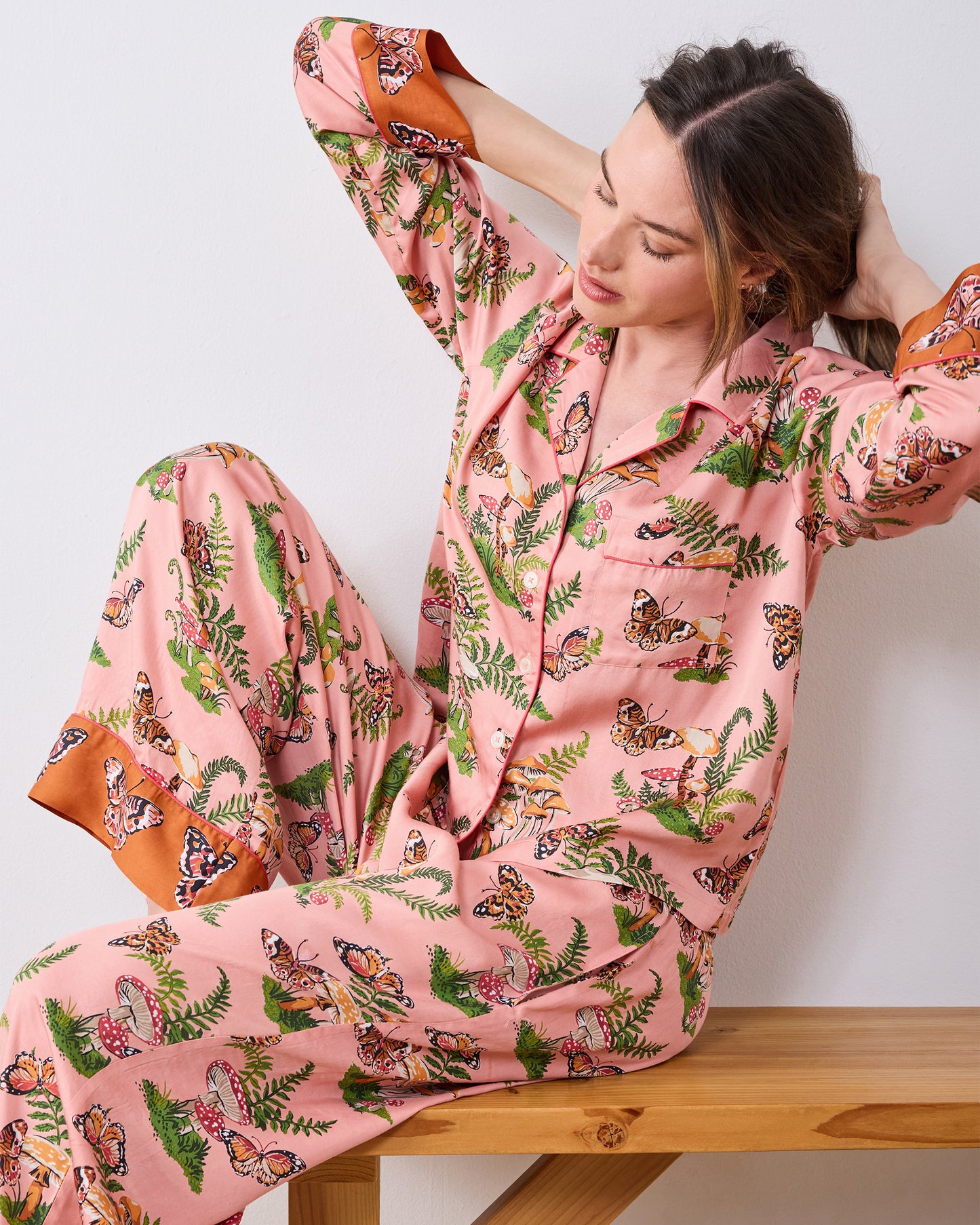 Person wearing the pink Butterfly Forager tall satin Soho pajama set with long sleeves, wide-leg pants, and contrasting orange cuffs, seated on a wooden bench.