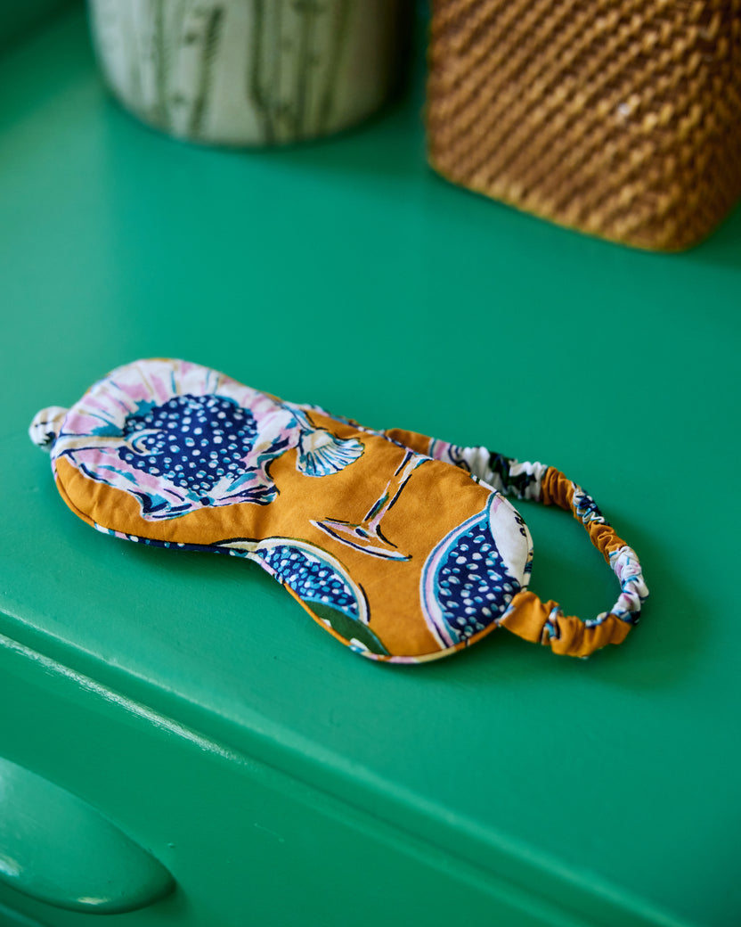 Goldenrod printed eye mask resting on a teal surface, with its elastic strap extended to the side.
