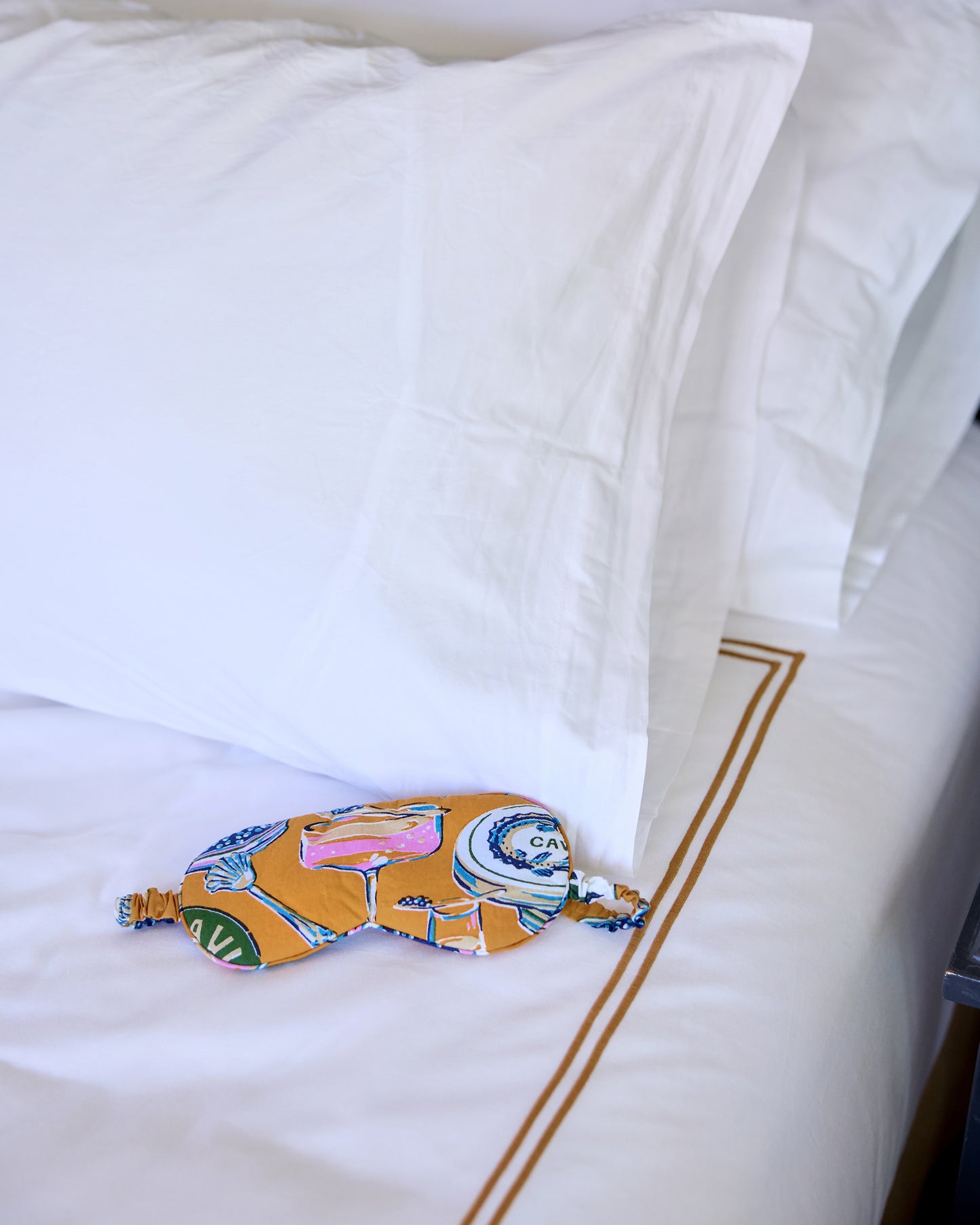 Goldenrod Champagne & Caviar eye mask resting on white bedding, with the elastic strap visible.