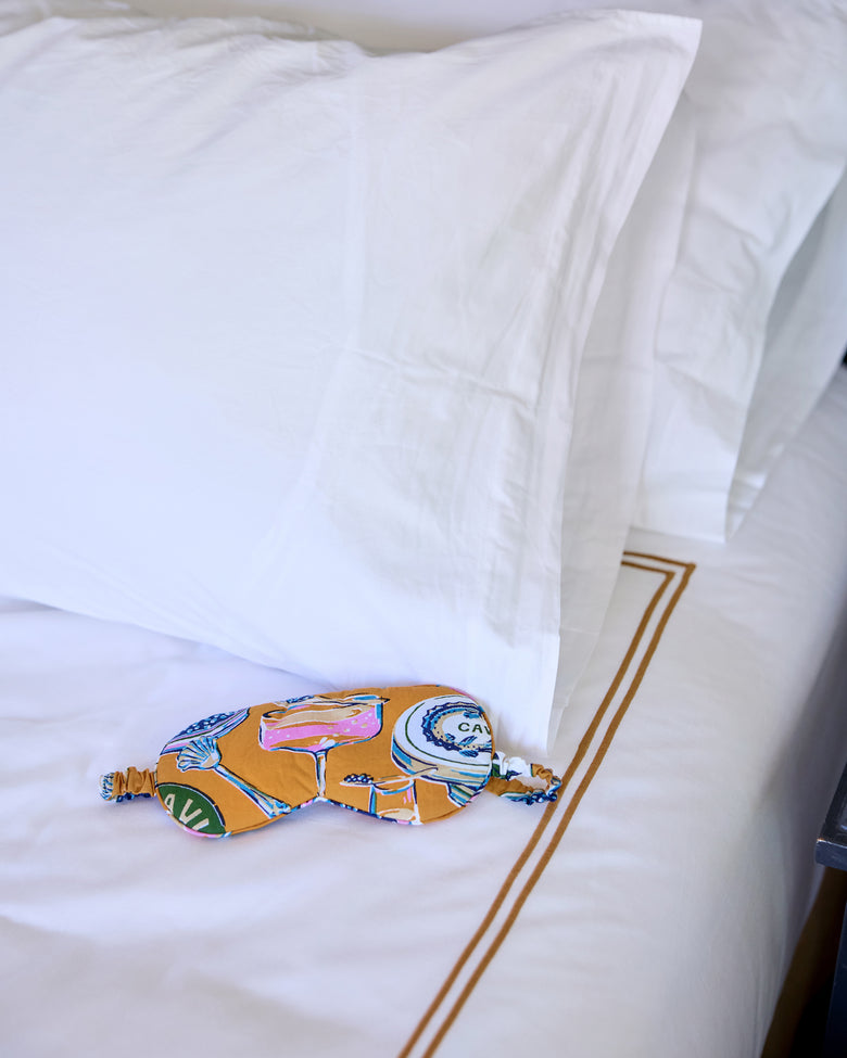 Goldenrod Champagne & Caviar eye mask resting on white bedding, with the elastic strap visible.