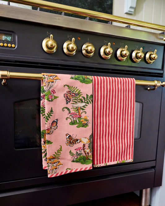 Two Butterfly Forager tea towels hanging from an oven handle, one showing the pink woodland print and the other showing red-and-white vertical stripes.