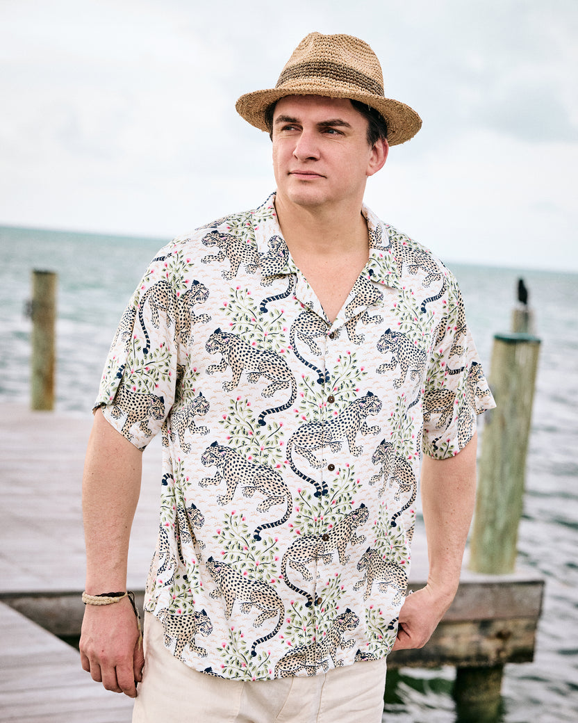 Model wearing the Wedding Cake Bagheera short-sleeve camp shirt with light trousers and a straw hat by the water
