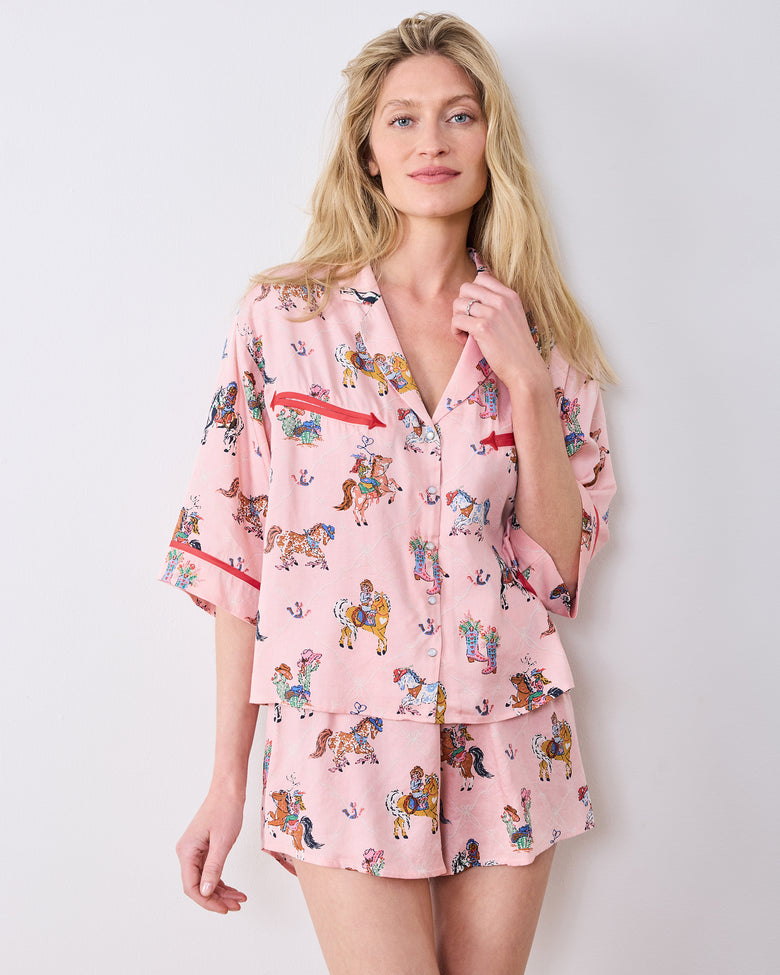 Model wearing the pink short pajama set with a horse-and-cowgirl print, collared top, red-trimmed chest pockets, and matching shorts.