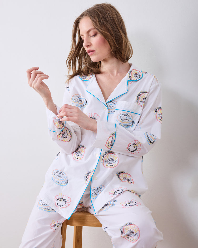 Model adjusting the sleeve of a white long-sleeve pajama set with embroidered caviar-tin motifs and blue piping.