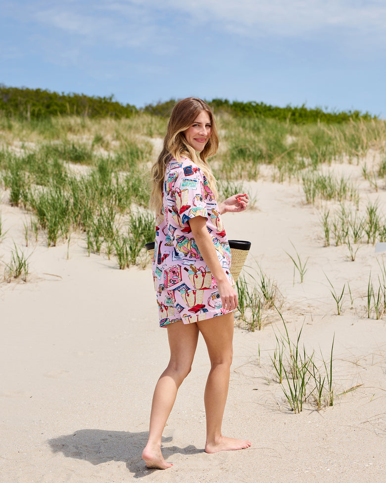 Side and back view of a person wearing a pink short-sleeve pajama set with a travel-themed print on a sandy beach.
