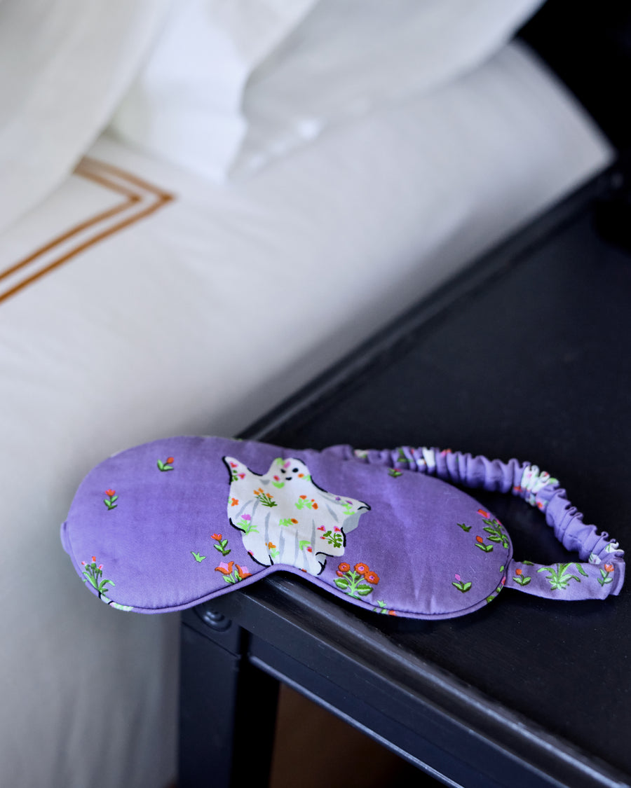 Purple eye mask with a white ghost appliqué resting on a dark surface beside a white pillow.