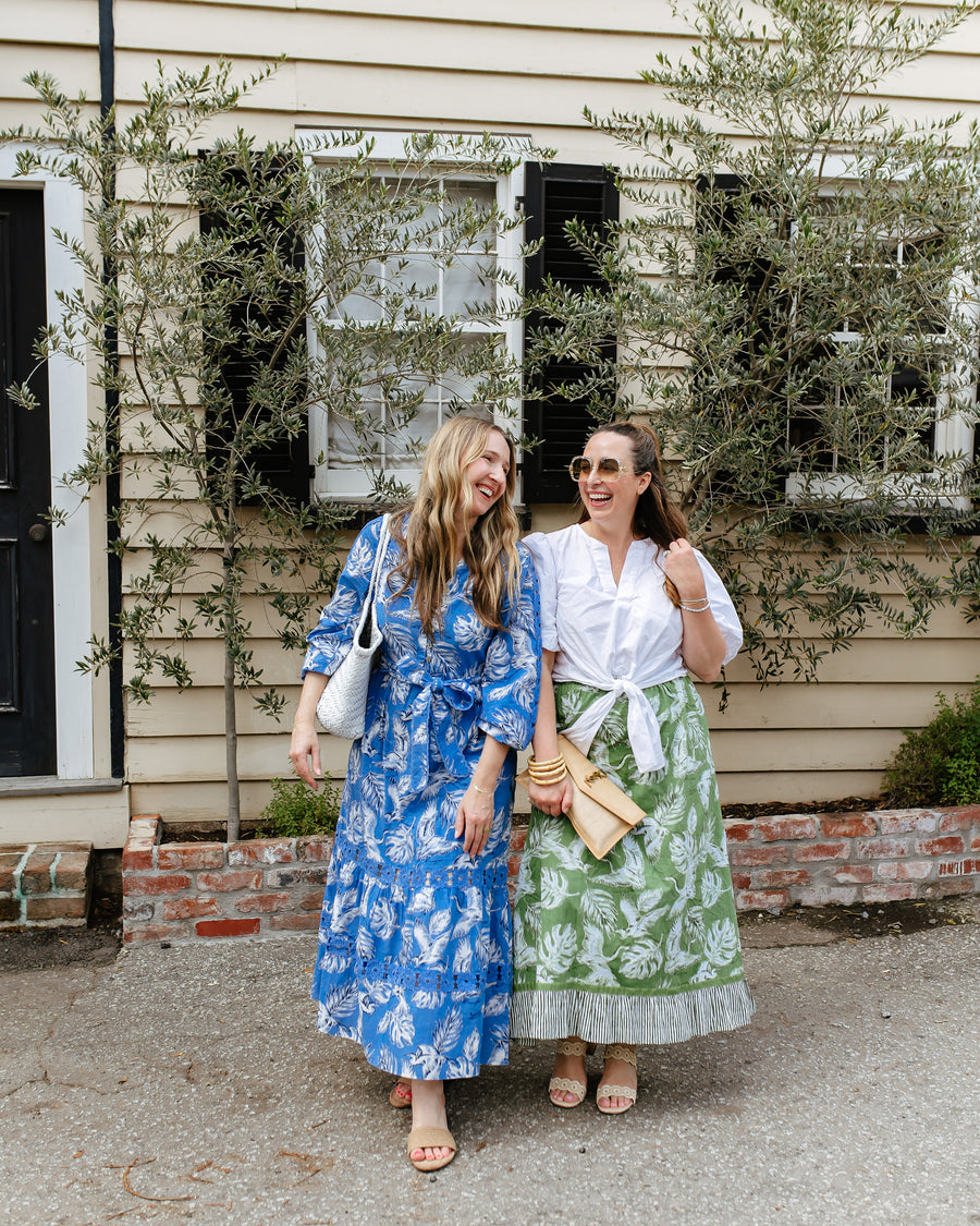 Two people outdoors, one wearing the blue Heron Landing Monroe dress and the other wearing a white top with a green patterned skirt.
