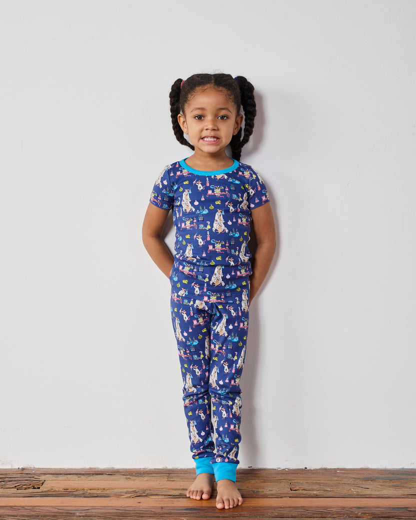 Child standing against a wall wearing the complete blue short-sleeve pajama set with a colorful laboratory-themed print.