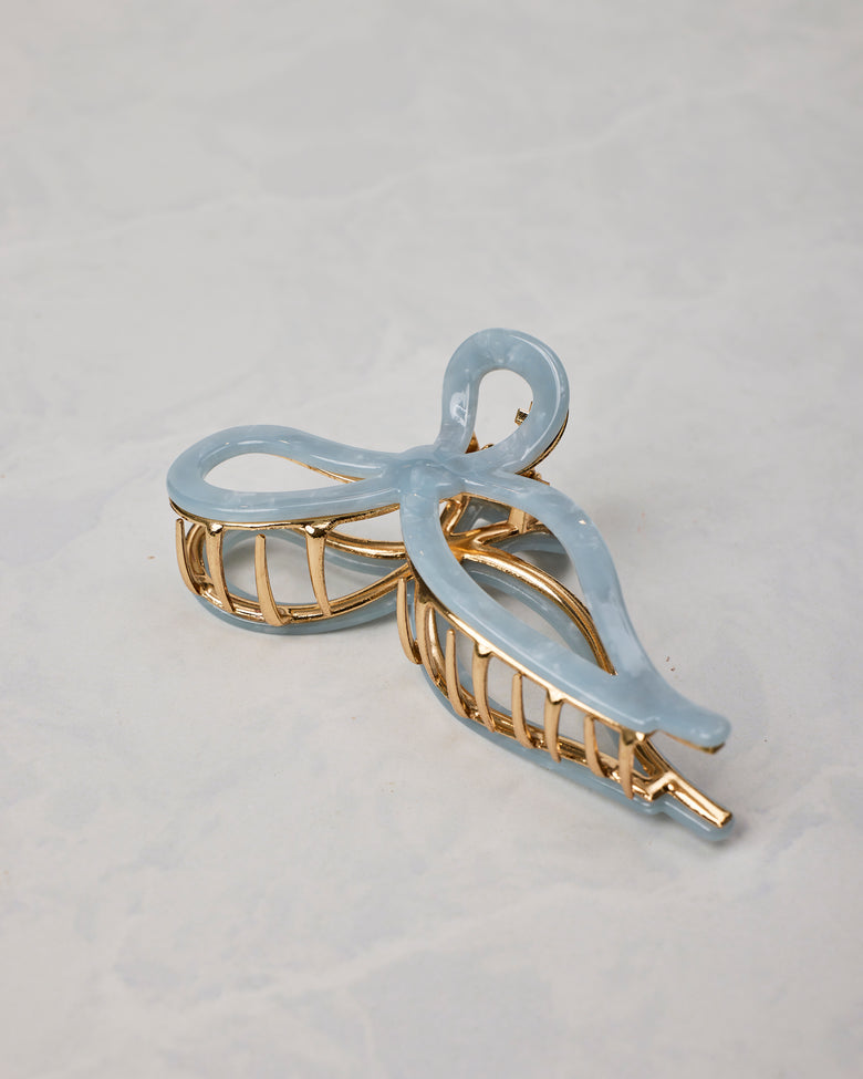 Pale blue ballet bow claw hair clip shown at an angle, with gold-toned teeth and frame.