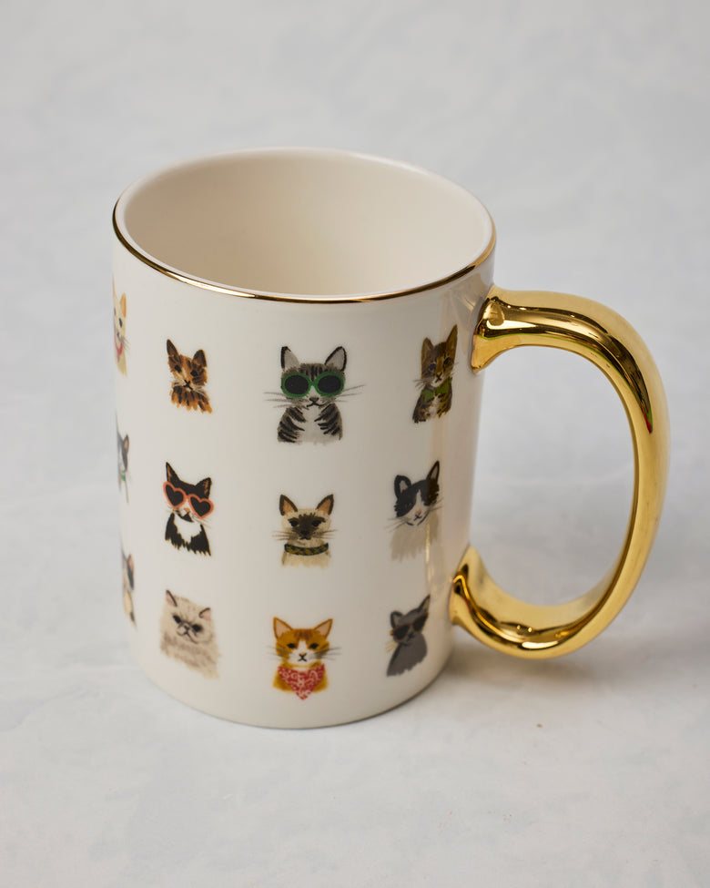 White mug with illustrated cats wearing accessories and a gold-colored handle and rim