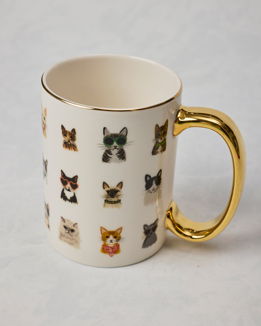 White mug with illustrated cats wearing accessories and a gold-colored handle and rim