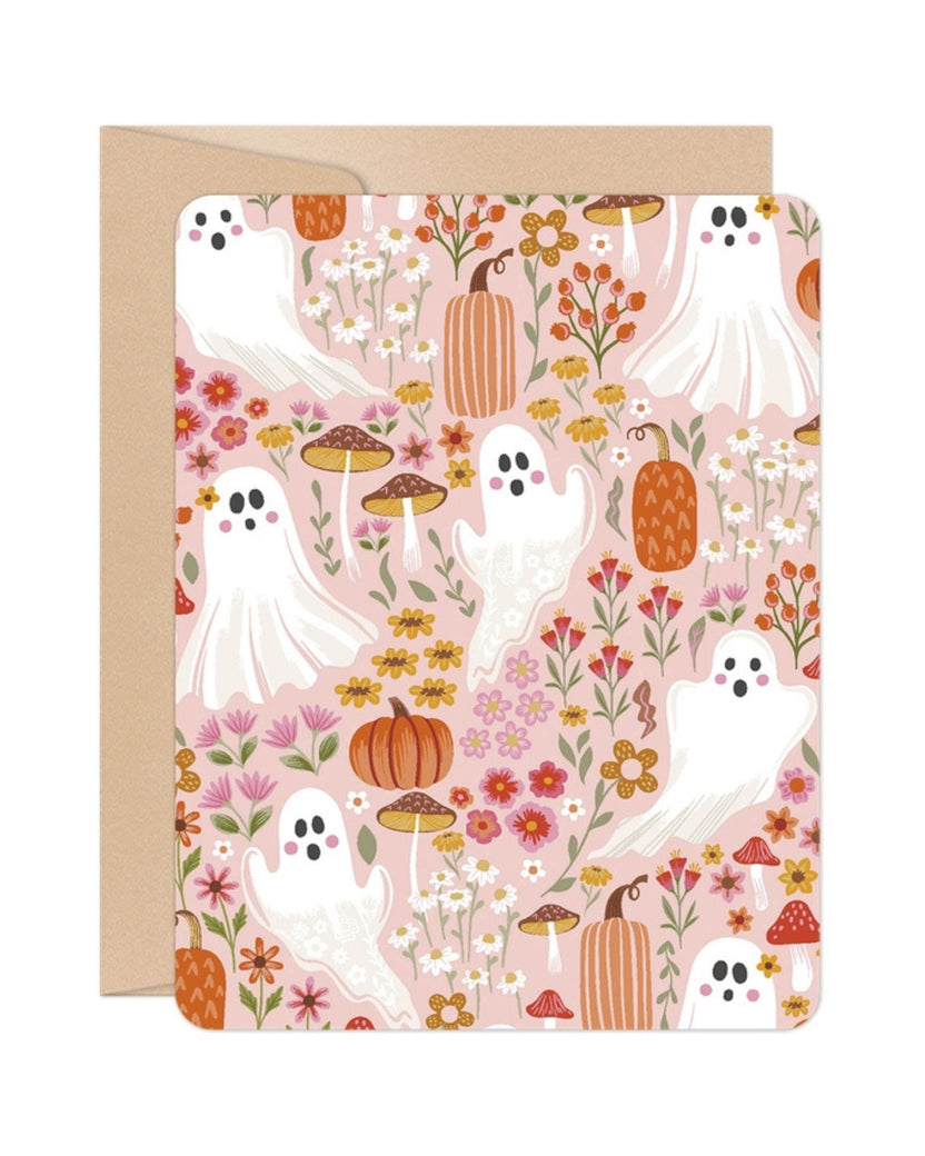 Front of a pink Halloween greeting card patterned with white ghosts, pumpkins, flowers, and mushrooms, shown with a kraft envelope.