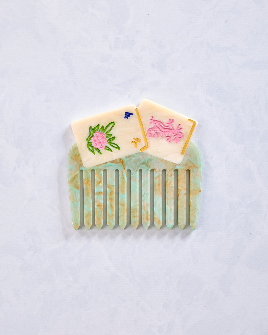 Green and cream decorative pocket comb with vertical teeth and painted floral and symbol details, shown on a light background.