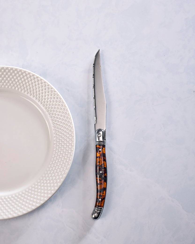 Single Laguiole steak knife with a serrated stainless steel blade and tortoise shell-style handle beside a white plate.