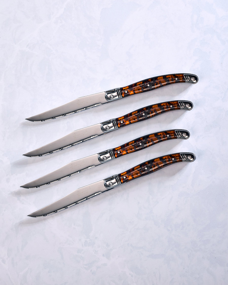 Four Laguiole steak knives with serrated blades and tortoise shell-style handles arranged diagonally.