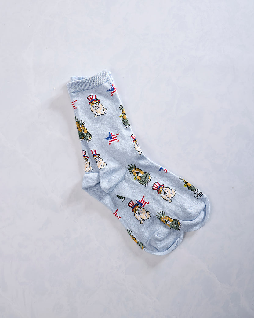 Pair of light blue crew socks with repeated patriotic dogs, pineapple-like figures, and American flag motifs.