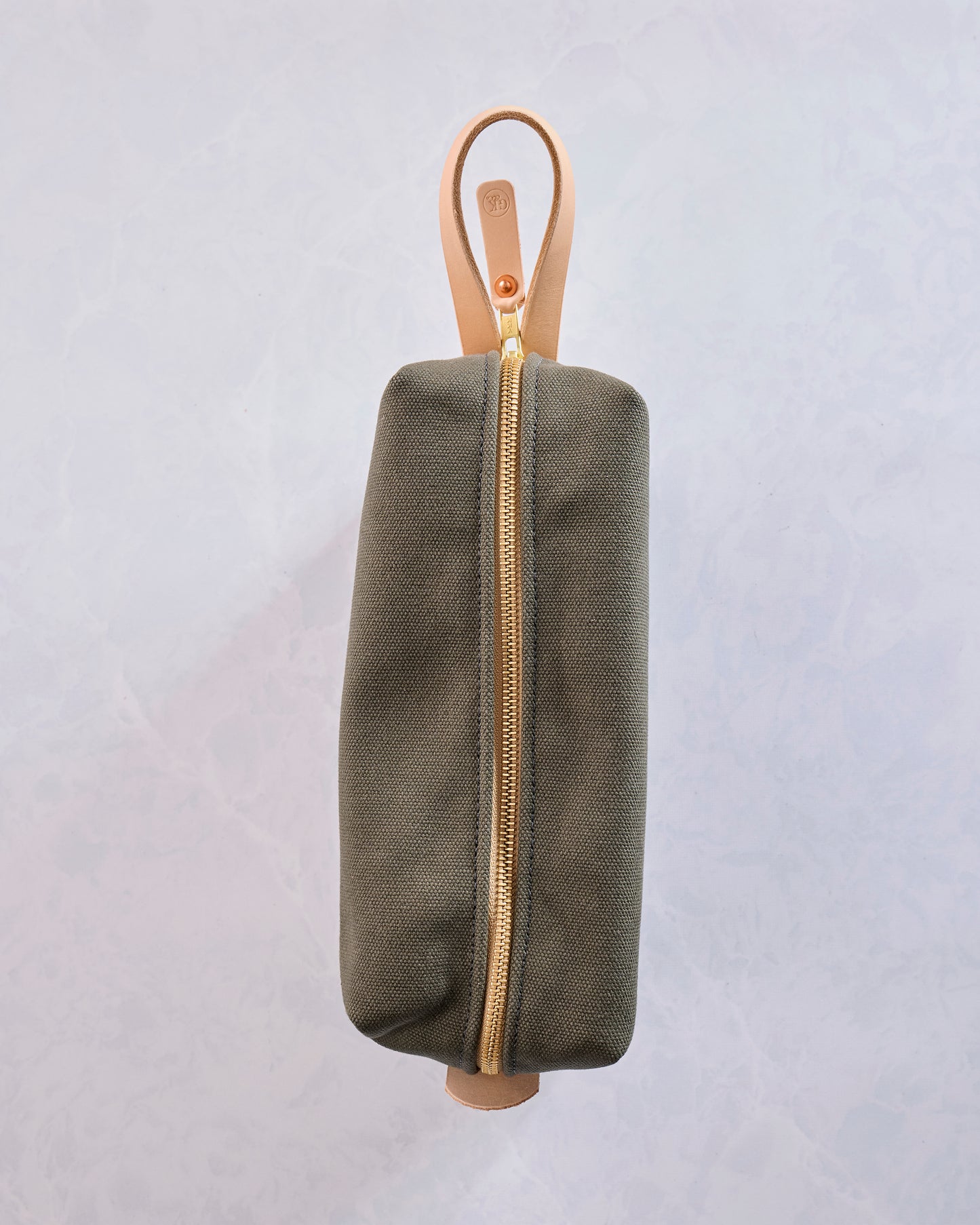 Side view of the olive canvas travel kit with a gold zipper, tan leather loop handle, and rounded rectangular shape.