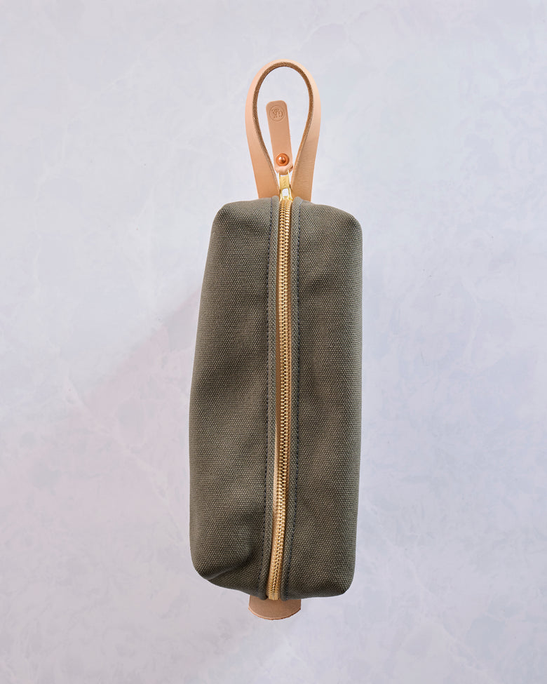 Side view of the olive canvas travel kit with a gold zipper, tan leather loop handle, and rounded rectangular shape.