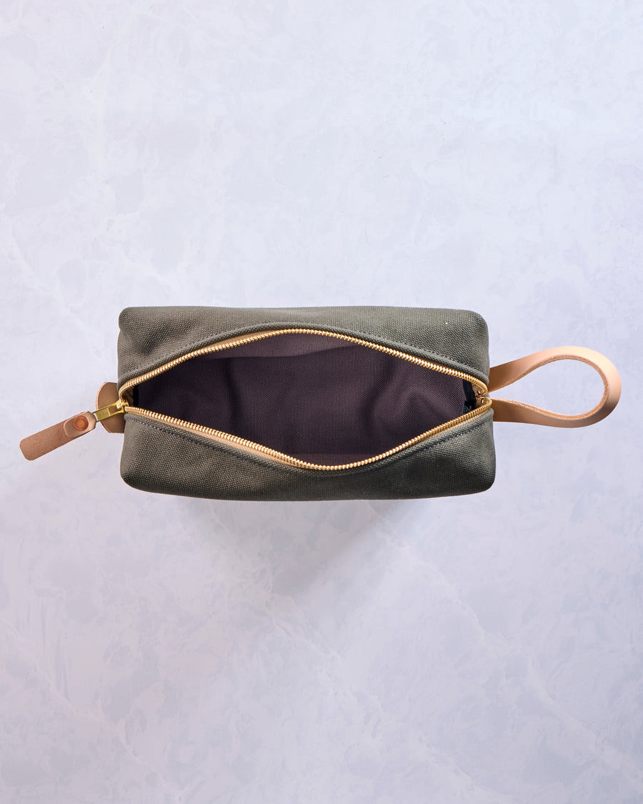 Olive canvas travel kit shown open, with a dark interior, gold-tone zipper, and leather wrist strap.