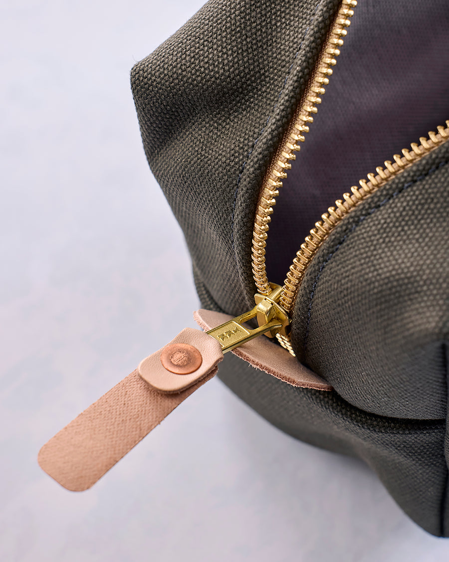 Close-up of the olive canvas travel kit's gold zipper, leather pull tab, and stitched seam.